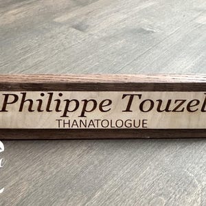 May include: A dark brown wooden desk nameplate with the name "Philippe Touzel" and the title "Thanatologue" engraved in a lighter wood.