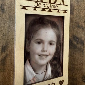 May include: A wooden photo frame with the word "DADA" at the top, and "Je t'aime" below it. A black and white photo of a young girl is in the center. The name "de Noah" is at the bottom with a heart symbol.