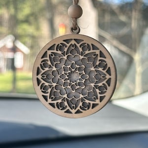 May include: A wooden car charm featuring a detailed mandala design. The charm is brown with a black mandala pattern and hangs from a brown string.