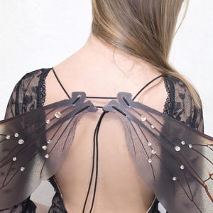 May include: A back view of a person wearing black lace clothing and a pair of translucent fairy wings. The wings are adorned with small, sparkling embellishments and have a delicate, veined pattern.