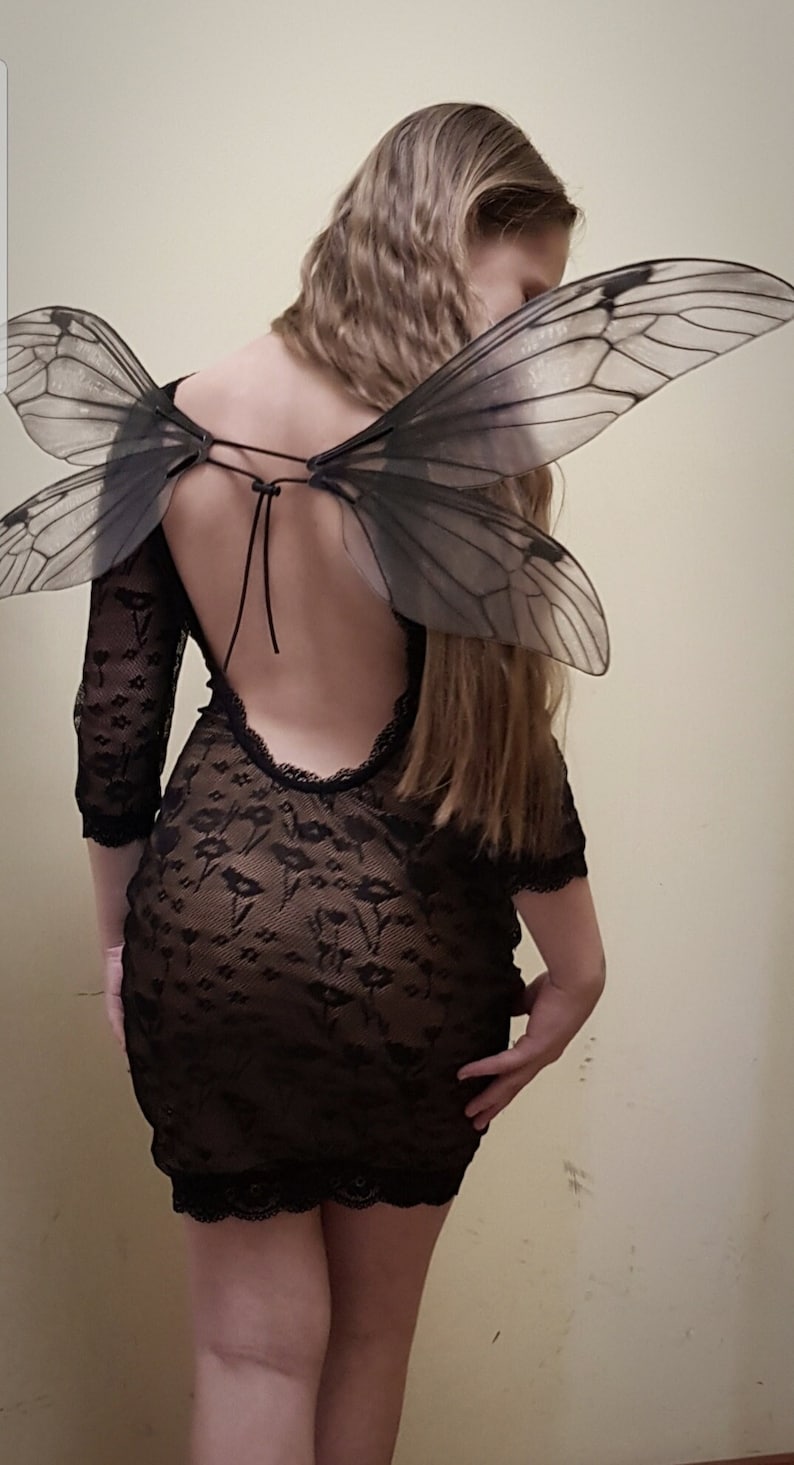 Puede incluir: Una mujer con un vestido negro de encaje con la espalda descubierta y un par de alas grandes, negras, parecidas a las de un insecto.