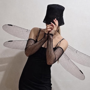 May include: A black spaghetti strap dress with a side slit, paired with black fishnet arm sleeves, a black crochet bucket hat, and large, translucent, black-veined dragonfly wings extending from the back of the garment.