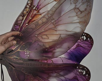 May include: A pair of translucent fairy wings featuring a gradient of purple and amber tones. The wings have a delicate, vein-like structure with intricate, bubbly textures and gold-colored accents throughout the surface. Black cords are attached.