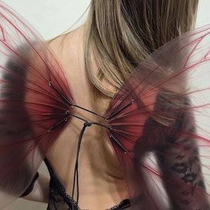 May include: Red and black fairy wings with a transparent design. The wings are attached with black string and are worn over a black lace top. The wings have a dark red gradient and are semi-transparent.