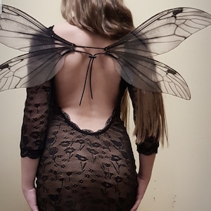 Puede incluir: Una mujer con un vestido negro de encaje con la espalda descubierta y un par de alas grandes, negras y transl&uacute;cidas.