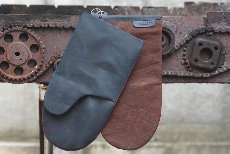 May include: Two oven mitts, one dark blue and one brown, hanging on a rusty metal chain and gear mechanism. The dark blue mitt has a leather texture and the brown mitt has a canvas texture. The brown mitt has the word "GOODAITY" printed on it.