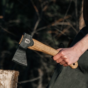Może przedstawiać: Topór z ciemną metalową głowicą i jasnym drewnianym trzonkiem. Na głowicy topora znajduje się wytłoczone logo. Trzonek trzyma osoba. Tło jest rozmyte, z widocznymi drzewami i liśćmi.