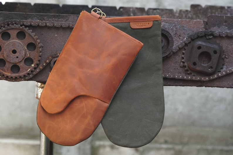 May include: A pair of oven mitts, one brown leather and one green canvas, hanging from a metal chain.