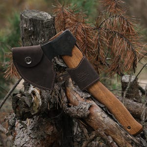 Puede incluir: Un hacha con mango de madera, cabeza de metal oscuro y funda de cuero marrón. El hacha descansa sobre un tocón y ramas, con un fondo de bosque. La funda de cuero tiene un cierre a presión.