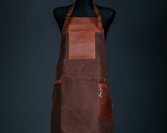 May include: A brown apron with a leather pocket and accents. The apron is displayed on a black mannequin. The apron has a large front pocket and a smaller pocket with a metal clasp. The leather pocket has a logo embossed on it.