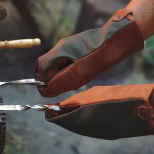 May include: A pair of brown and green leather grilling gloves with a strap on the wrist. The gloves are being used to hold a metal skewer with food on it over a fire.