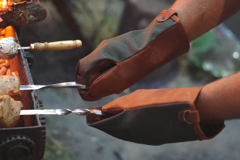 May include: A pair of brown and green leather grilling gloves with a strap on the wrist. The gloves are being used to hold a metal skewer with food on it over a fire.