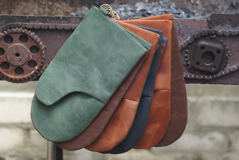 May include: A set of five leather oven mitts in various colors, including green, brown, orange, navy blue, and tan. The mitts are stacked on top of each other and are hanging from a metal chain.