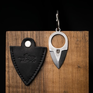 Könnte beinhalten: Ein kleines, silberfarbenes Messer mit schwarzer Klinge und Schlüsselring. Daneben befindet sich eine schwarze Lederscheide mit einem dekorativen Emblem. Der Hintergrund ist eine Holzoberfläche.