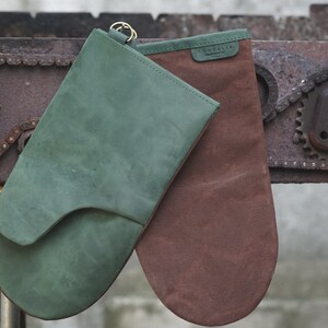 May include: Two oven mitts, one green and one brown, hanging on a rusty metal surface. The green mitt has a loop for hanging.