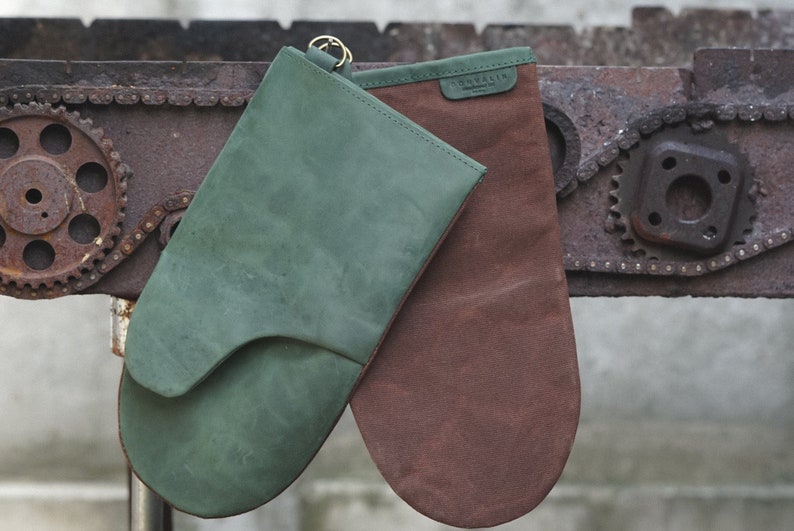 May include: Two oven mitts, one green and one brown, hanging on a rusty metal surface. The green mitt has a loop for hanging.