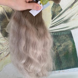 Puede incluir: Un paquete de extensiones de cabello humano ondulado, rubio claro. El cabello está atado con una banda elástica verde y tiene una pequeña etiqueta blanca con el número "12" escrito en ella.