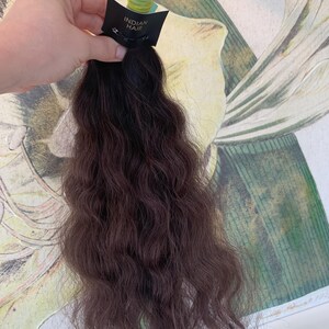 Puede incluir: Un paquete de extensiones de cabello humano ondulado marrón oscuro con una etiqueta negra que dice "INDIAN HAIR".