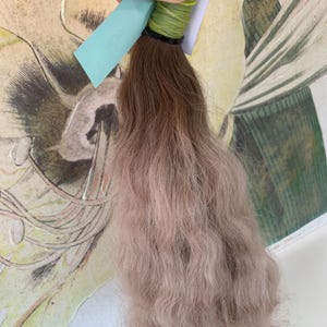 May include: A bundle of wavy human hair extensions with a light brown to blonde ombre color. The hair is tied with a green elastic band and a blue tag.