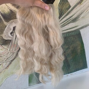 May include: A bundle of wavy, platinum blonde human hair extensions. The hair is tied together with a black elastic band and has a tag that reads "Indian Hair".