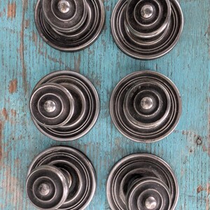 May include: Six antique silver-toned cabinet knobs with a concentric circle design. Each knob features a central, raised button surrounded by layered, circular ridges. The knobs are arranged on a distressed, turquoise-painted wooden surface.