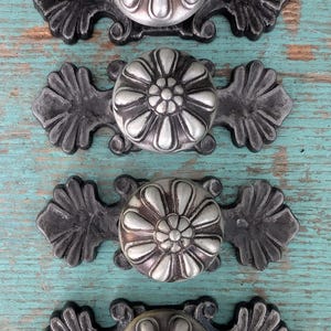May include: Four decorative cabinet knobs with a floral design. The knobs are silver-colored with a dark gray base and mounting hardware. The background is a distressed turquoise-colored wooden surface.