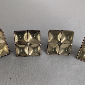 May include: Four square-shaped metal cabinet knobs with a metallic gold finish. Each knob features a grid pattern consisting of four concave, rounded square indentations arranged in a two-by-two formation, creating a textured, geometric surface.