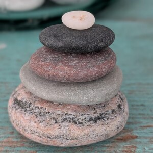 May include: A stack of smooth, colorful stones arranged in a balanced tower. The stones vary in size and color, ranging from light pink to dark gray. A small, white stone sits atop the stack, creating a sense of harmony and balance. The arrangement is set against a turquoise background.