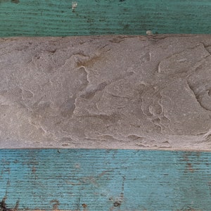 May include: A gray, rectangular stone with a rough, textured surface. The stone has a slightly curved shape and is sitting on a blue wooden surface.