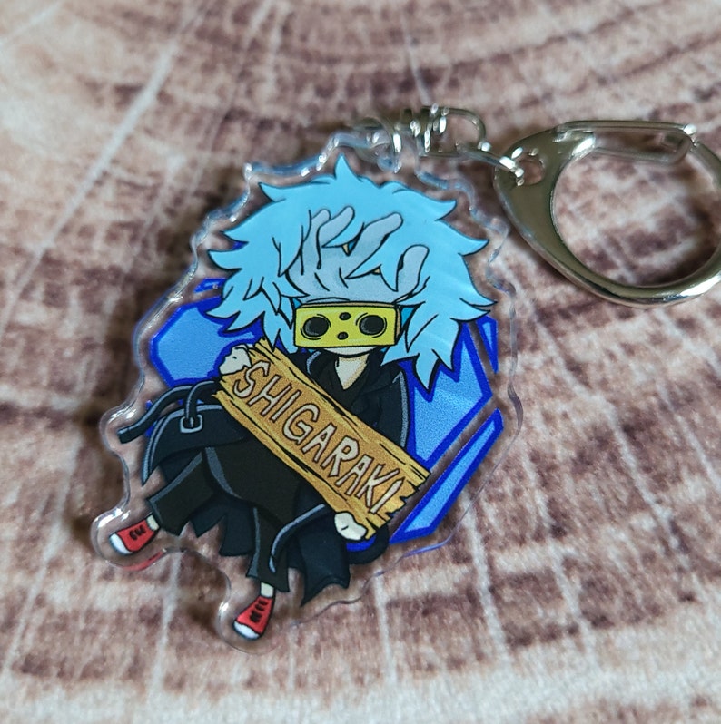 May include: A blue and black anime character keychain with the text "SHIGARAKI" on a wooden sign. The character has blue hair and is wearing a black mask with yellow eyes.