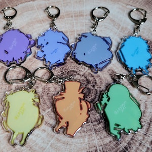 May include: Set of 7 colorful acrylic keychains featuring various cartoon characters. Each keychain has a different design and includes the text "@lastam_art".
