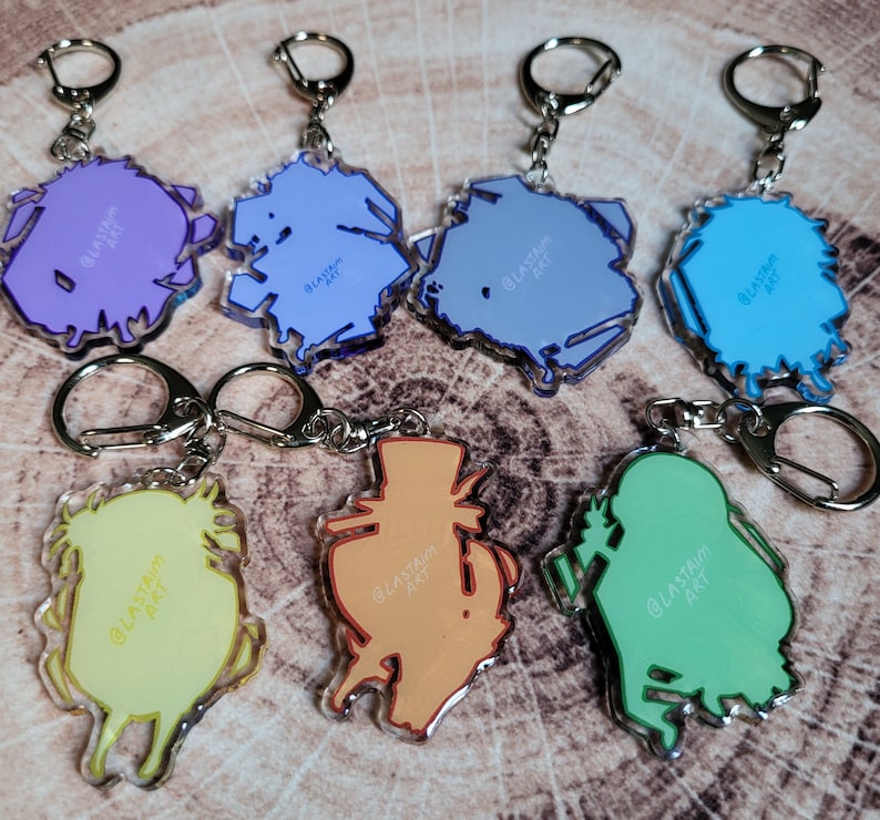 May include: Set of 7 colorful acrylic keychains featuring various cartoon characters. Each keychain has a different design and includes the text "@lastam_art".