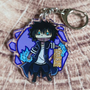 May include: A blue and purple anime character keychain with blue flames and the word "DABI" on a wooden sign. The character has black hair, blue eyes, and a white shirt.