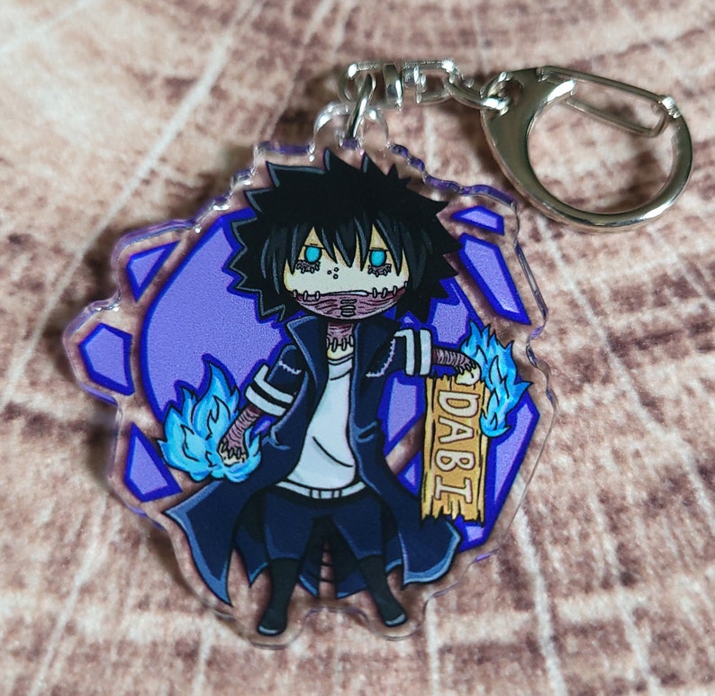 May include: A blue and purple anime character keychain with blue flames and the word "DABI" on a wooden sign. The character has black hair, blue eyes, and a white shirt.