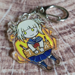 May include: A clear acrylic keychain featuring a cartoon character with white hair, yellow eyes, and a blue and white sailor uniform. The character is holding a wooden sign that says "TRA".