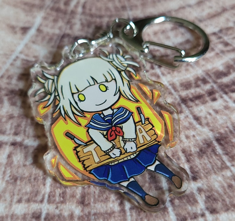 May include: A clear acrylic keychain featuring a cartoon character with white hair, yellow eyes, and a blue and white sailor uniform. The character is holding a wooden sign that says "TRA".