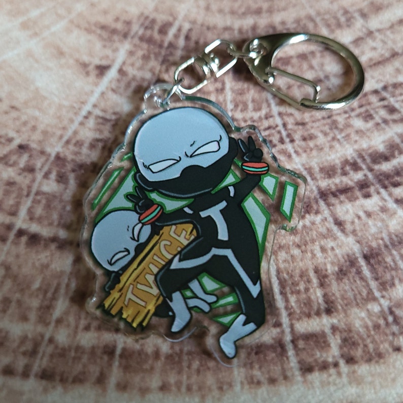 May include: A black and white anime character keychain with green and white accents. The character is holding a smaller version of themself with the word "TWICE" written on a wooden sign.