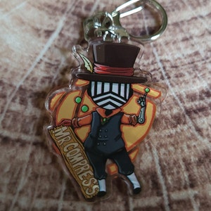 May include: A clear acrylic keychain featuring a cartoon character with a black top hat, a black and white mask, and a yellow and orange background. The character is holding a wooden sign that says "MR. COMPRESS".
