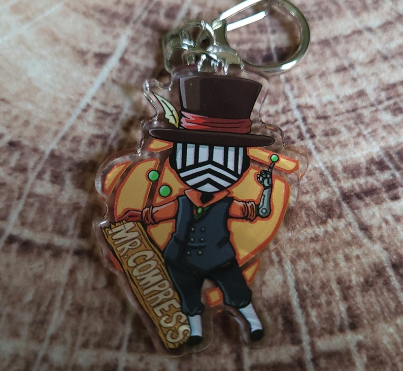 May include: A clear acrylic keychain featuring a cartoon character with a black top hat, a black and white mask, and a yellow and orange background. The character is holding a wooden sign that says "MR. COMPRESS".