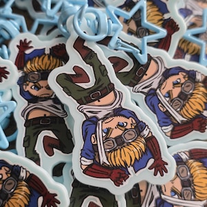 May include: A collection of anime-style keychains featuring a character in a blue jacket, goggles, and green pants. The keychains have a light blue border and a matching star-shaped clasp.