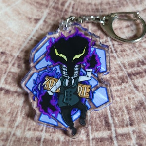 May include: A purple and black anime character keychain with a dark purple flame design. The character is holding a wooden sign that says "ZUKURI".