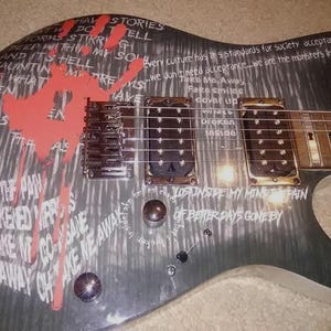 May include: An electric guitar body featuring a grey and black brushed finish with white printed text lyrics and a prominent red handprint graphic. The instrument includes two black humbucker pickups, a bridge, and two metallic control knobs.