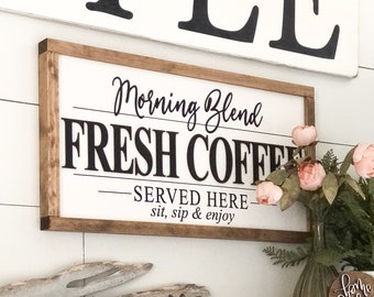 May include: A wooden sign with the text "Morning Blend Fresh Coffee Served Here sit, sip & enjoy" hanging on a white wall. A wooden shelf with a "Welcome" sign and a mug rack with various mugs hanging on it.