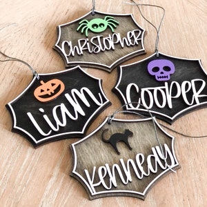 K&ouml;nnte beinhalten: Vier h&ouml;lzerne Halloween-Anh&auml;nger in Form von Spinnweben mit wei&szlig;em Rand. Jeder Anh&auml;nger ist mit einem anderen Design verziert: einer Spinne, einem Sch&auml;del, einem K&uuml;rbis und einer schwarzen Katze. Die Anh&auml;nger sind mit den Namen Christopher, Cooper, Liam und Kennedy personalisiert.