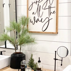 May include: A bathroom vanity with a black and white sign that reads "Jingle all the way". There is a black vase with a green plant, two clear glass bottles with black labels that say "Hand Soap", a small green pine tree, and a white candle on a black tray. There is also a black magnifying mirror on a stand.