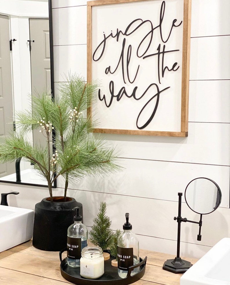 May include: A bathroom vanity with a black and white sign that reads "Jingle all the way". There is a black vase with a green plant, two clear glass bottles with black labels that say "Hand Soap", a small green pine tree, and a white candle on a black tray. There is also a black magnifying mirror on a stand.