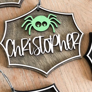 K&ouml;nnte beinhalten: Ein h&ouml;lzerner Halloween-Anh&auml;nger in Form eines Spinnennetzes mit einer gr&uuml;nen Spinne in der Mitte. Der Anh&auml;nger ist braun lackiert und hat einen wei&szlig;en Rand. Der Name "Christopher" ist in wei&szlig;en Kursivbuchstaben auf dem Anh&auml;nger geschrieben.