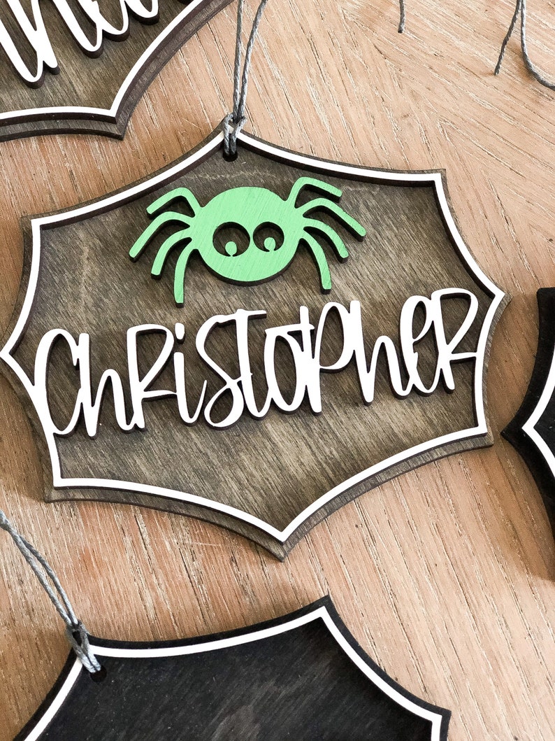 K&ouml;nnte beinhalten: Ein h&ouml;lzerner Halloween-Anh&auml;nger in Form eines Spinnennetzes mit einer gr&uuml;nen Spinne in der Mitte. Der Anh&auml;nger ist braun lackiert und hat einen wei&szlig;en Rand. Der Name "Christopher" ist in wei&szlig;en Kursivbuchstaben auf dem Anh&auml;nger geschrieben.