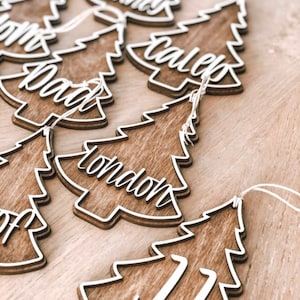 Peut inclure: Décorations de Noël en bois en forme d'arbre avec des lettres blanches. Les décorations sont décorées avec les noms "London", "Caleb", "Mommy", "Daddy", et "JJ".