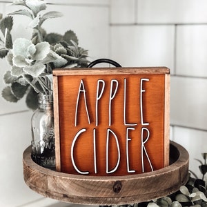 May include: A wooden sign with a rustic orange background and white lettering that reads "APPLE CIDER". The sign is sitting on a tiered wooden tray with greenery.